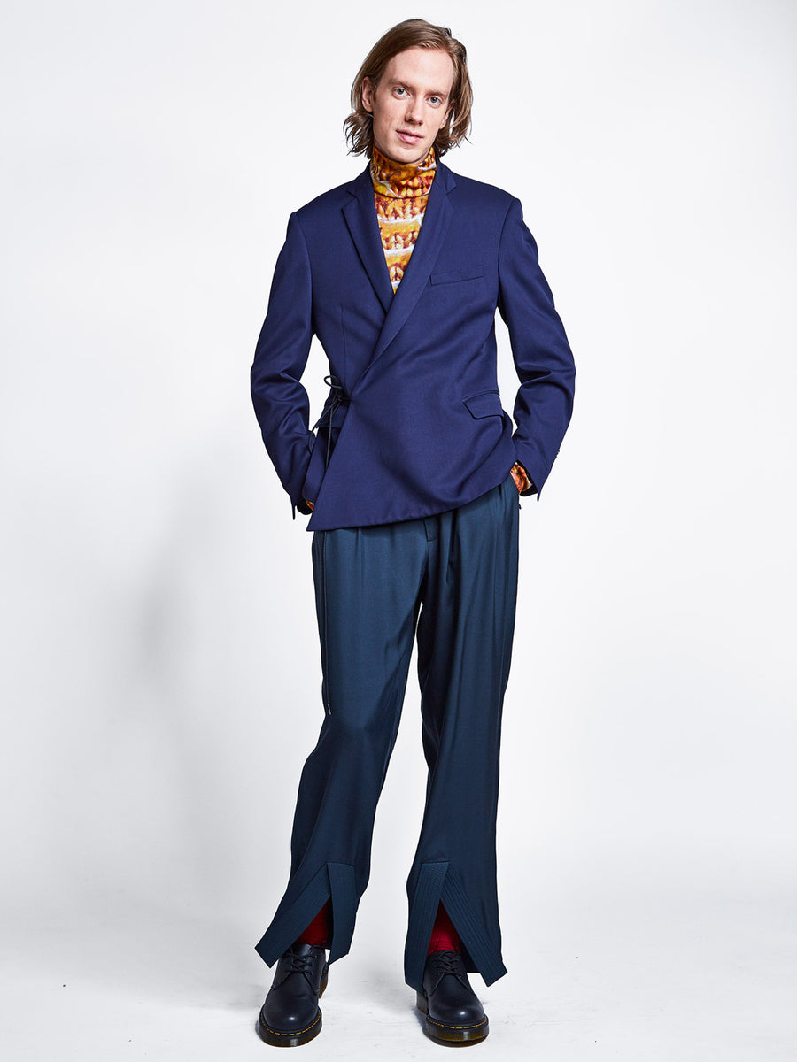Men's navy blue tailored wrap blazer jacket with cord tie over teal pleated wide leg pants and printed turtleneck.